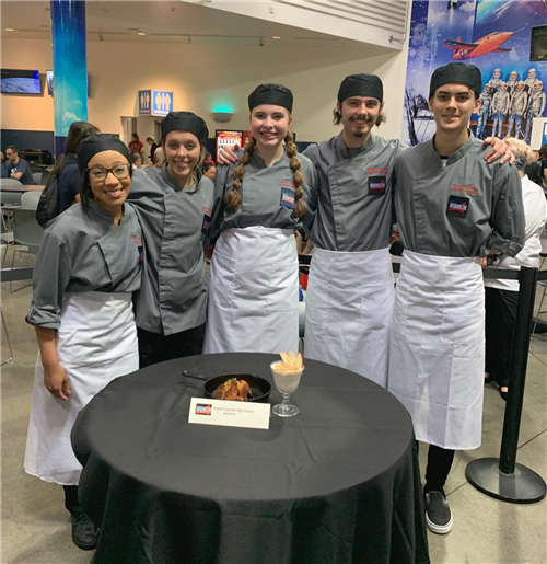 culinary students - five - in HT cooking uniforms with NASA badges and white aprons, smiling for group photo with food